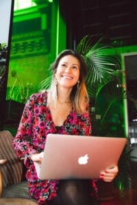 Smiling woman using a laptop in a modern indoor setting with lush greenery.