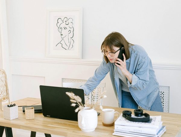 Woman multitasking with laptop and phone in modern home office setting.
